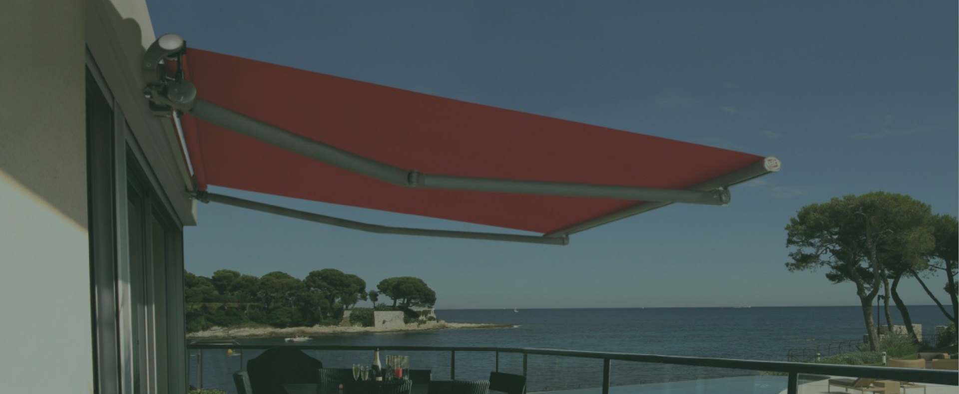 Tenda da sole rossa posizionata sopra un balcone con vista sul mare, su uno sfondo di alberi e cielo sereno.