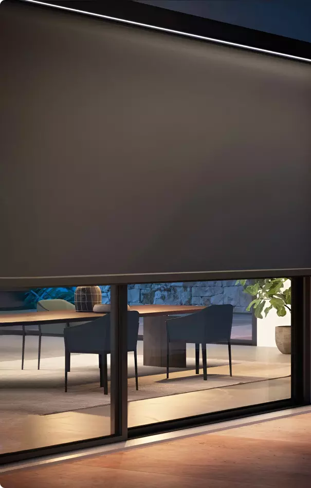 Vista parziale di una sala da pranzo con una grande finestra. Si notano un tavolo di legno e sedie scure. Dietro, si intravede una pianta e una parete in pietra.